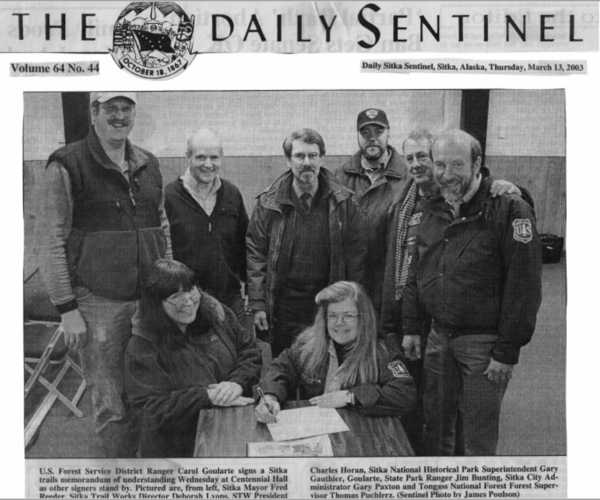 A newspaper photo from the Daily Sitka Sentinel from Thursday, March 13, 2002. This photo shows an assortment of representatives from the US Forest Service, Sitka Trail Works, Sitka National Historical Park, Alaska State Parks, and Sitka City Government officials, etc. signing a Sitka trails memorandum of understanding.