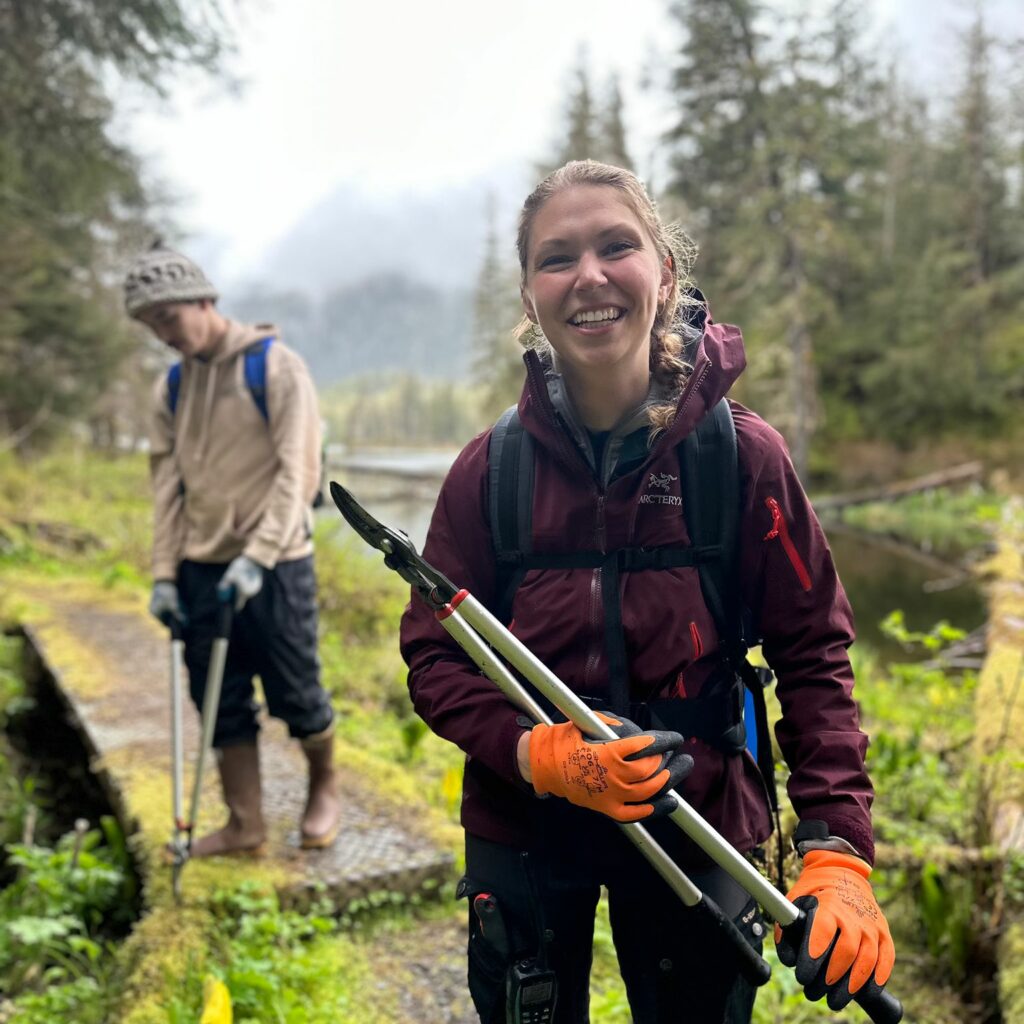 A photo of Emily Pound (she/they) smiling, holding a pair of loppers while doing trail work.