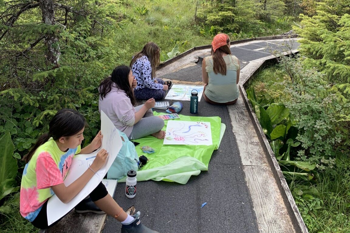 Youth in Sitka Trail Works' youth programming sit on a trail, coloring.