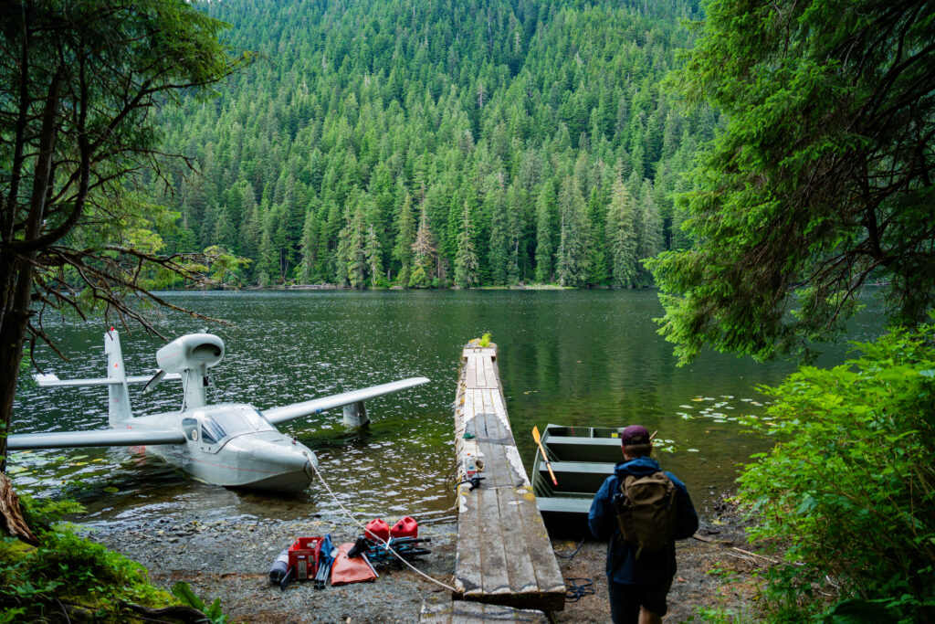 A hiker approaches salmon lake, with a float plane and a rowboat tied to either side of a dock ahead of them.